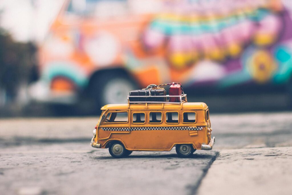 Close-up of a toy Volkswagen van with luggage, set against a colorful street backdrop, evoking summer travel vibes.